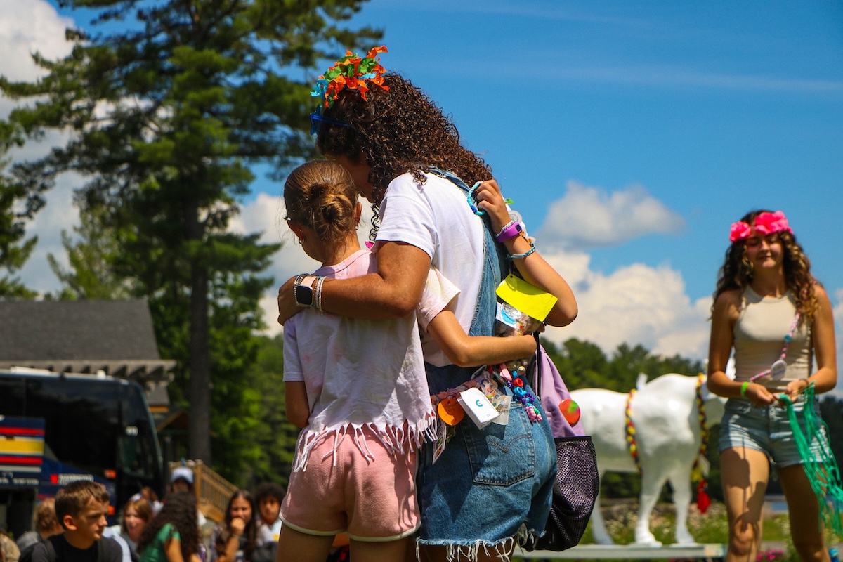 camper and counsellor with arms around each other, surrounded by others on a sunny day at camp