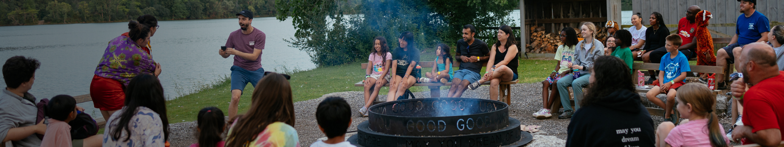 families and counsellors around the campfire at camp with lake in background