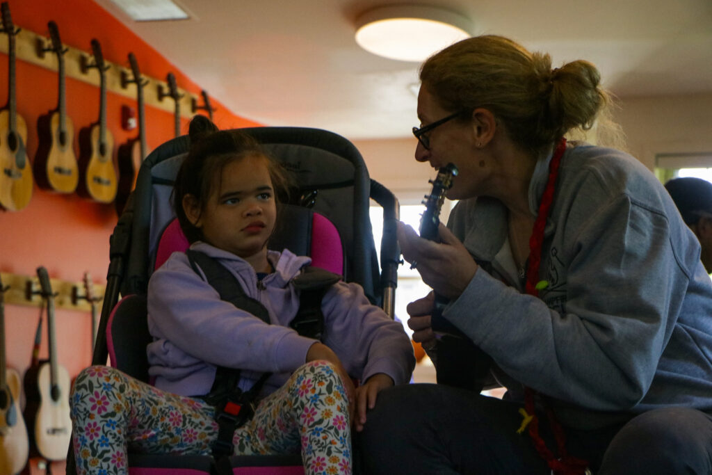 shaindy playing ukulele to camper in a wheelchair in the music room at camp