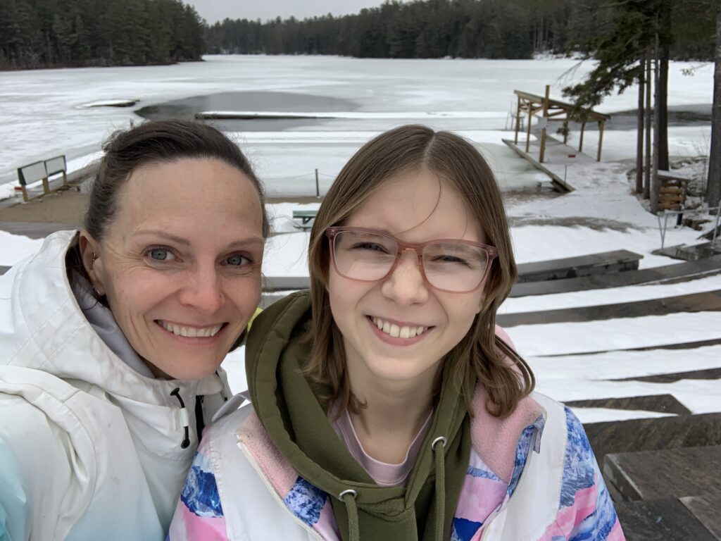 emma and mom at camp on a winter day with frozen lake and trees in the background