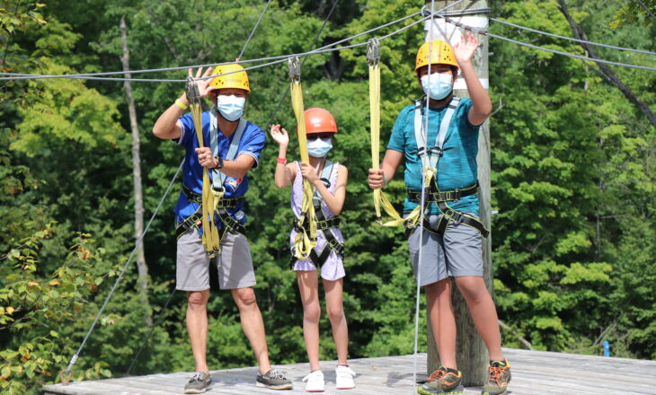 campers on adventure course at camp wearing protective helmets and harnesses with trees in the background