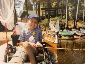 camper in wheelchair on dock with fishing rod and canoe and sailboats in background