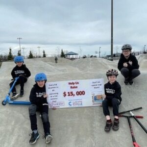 The Fundraiser Warriors. Four kids wearing helmets at skateboard park holding a giant cheque